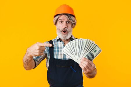 Picture of shocked elderly grey-haired bearded man builder in helmet posing isolated over yellow wall background holding money pointing.の写真素材