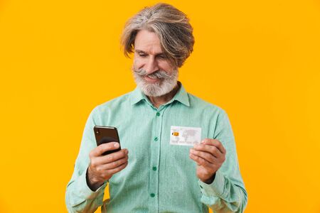 Photo of happy positive grey-haired bearded man in blue shirt posing isolated over yellow wall background using mobile phone holding credit card.の写真素材