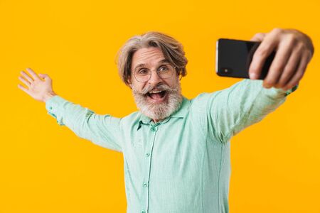 Image of happy emotional grey-haired man posing isolated over yellow wall background take a selfie by mobile phone.の写真素材