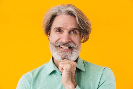 Image of positive happy elderly grey-haired bearded man in blue shirt posing isolated over yellow...