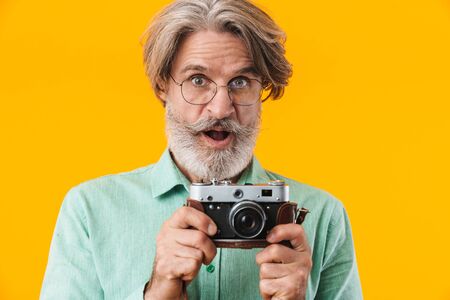 Image of shocked grey-haired man photographer posing isolated over yellow wall background holding camera.の写真素材