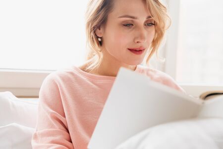 Image of a concentrated young woman indoors at home lies in bed under blanket reading book.