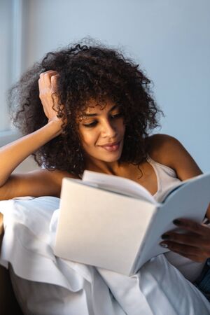 Image of a concentrated beautiful young african woman indoors at home reading book lies in bed.