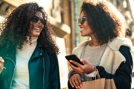 Two beautiful young stylish african women walking together at the street, using mobile phone,...
