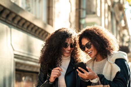 Two beautiful young stylish african women walking together at the street, using mobile phone, talkingの写真素材