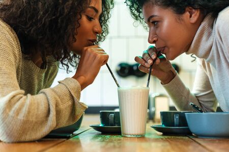 Two cheerful young african girlfriends sitting at the cafe drinking coffee, drinking one milkshake togetherの写真素材