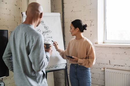 Image of multiethnic young female and male coworkers standing by flipchart while working in officeの写真素材