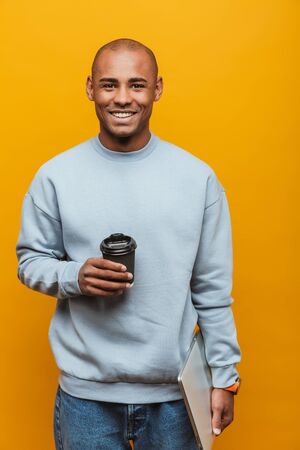 Portrait of an attractive smiling confident casual young african man standing over yellow background, carrying laptop computer, drinking takeaway coffeeの写真素材