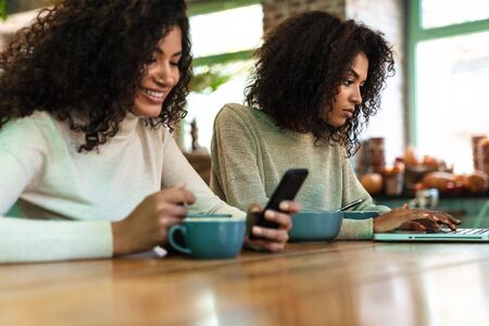 Two cheerful young african girlfriends sitting at the cafe drinking coffee, using mobile phone and...