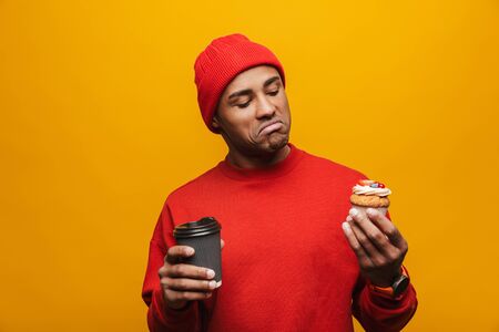 Portrait of an attractive confident casual young african man standing over yellow background, holding takeaway coffee cup and cupcakeの写真素材