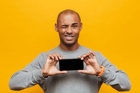 Portrait of an attractive smiling confident casual young african man standing over yellow background, showing blank screen mobile phoneの写真素材