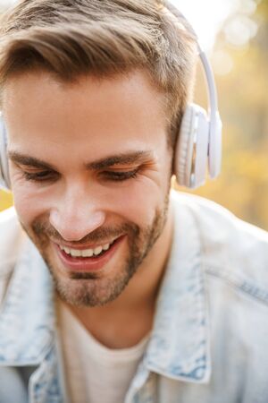 Image of handsome young caucasian man wearing headphones sitting on bench while walking in autumn parkの写真素材