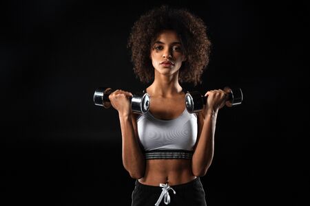 Image of confident african american sportswoman doing exercise with dumbbells isolated over black backgroundの写真素材