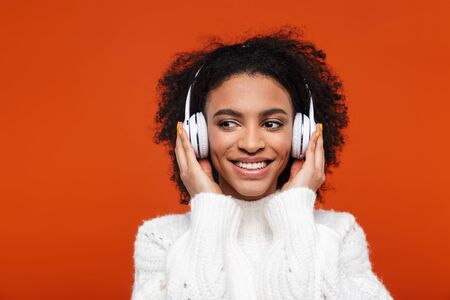 Cheerful young african woman listening to music with wireless headphones isolated over red backgroundの写真素材