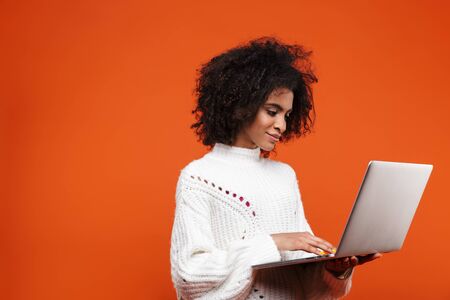 Attractive smiling young african woman standing isolated over red background, using laptop computerの写真素材