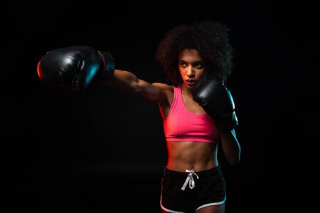 Image of young african american sportswoman working out in boxing gloves isolated over black backgroundの写真素材