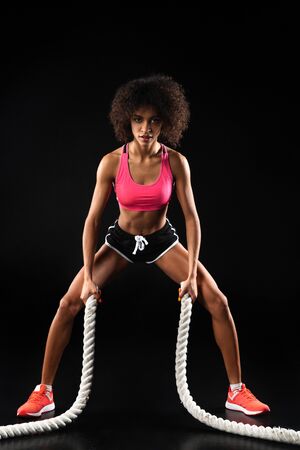 Image of athletic african american sportswoman doing exercise with battle ropes isolated over black backgroundの写真素材