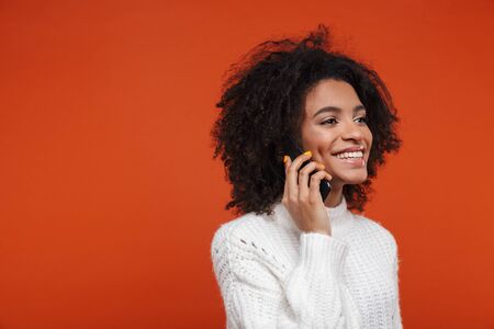 Attractive young african woman wearing sweater talking on mobile phone isolated over red backgroundの写真素材
