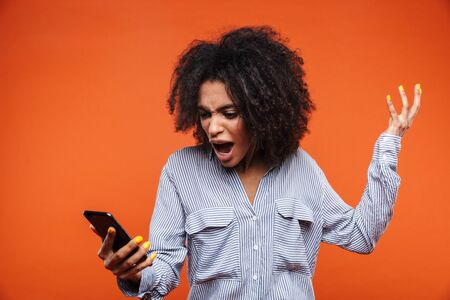 Angry young african woman talking on mobile phone isolated over red background, screamingの写真素材