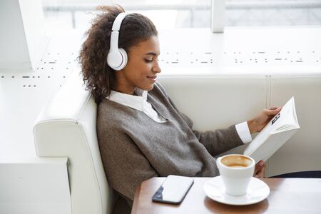 Portrait of young beautiful african american woman wearing earphones reading book while sitting on sofa in cafeの写真素材