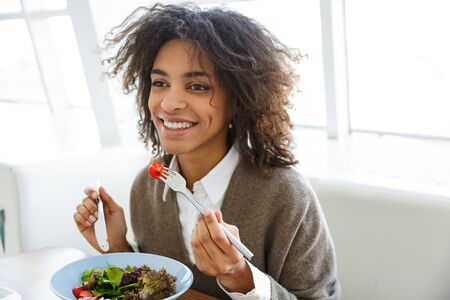 Portrait of young beautiful african american woman eating salad while having lunch in cafeの写真素材