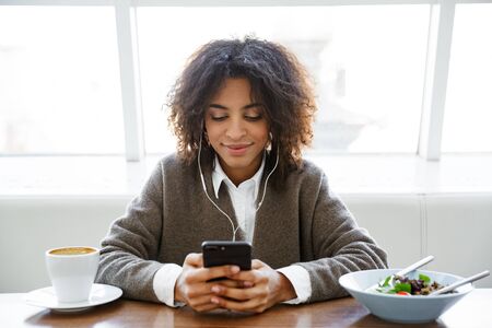 Portrait of young african american woman listening to music with earphones and cellphone in cafe