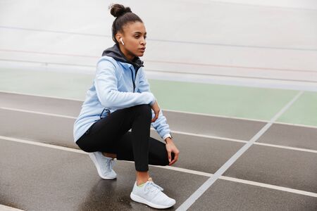 Attractive young african sportswoman resting after running at the stadium, wearing wireless earphonesの写真素材