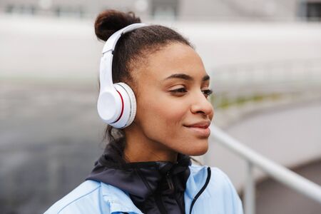 Attractive young healthy african sportswoman listening to music with wireless earphones wrking out...