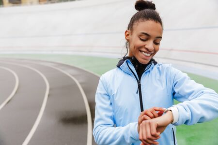 Attractive young african sportswoman resting after running at the stadium, looking at smartwatch