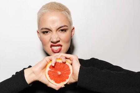 Portrait of an attractive happy young blonde short haired woman showing halved grapefruit isolated...