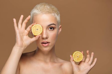 Image of a serious young blonde stylish woman with short haircut posing isolated over dark beige wall background covering eyes with lemon.の写真素材