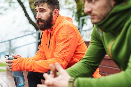 Close up of two fit young sportsmen resting after work out outdoors at the playground, holding water...