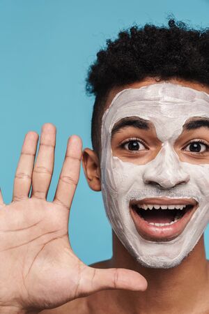 Photo of joyful african american man in facial cosmetic mask showing his palm isolated over blue backgroundの写真素材