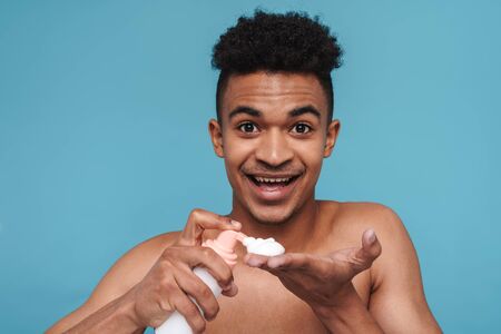 Photo of shirtless african american man smiling while applying facial cleanser foam isolated over blue backgroundの写真素材