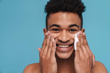 Photo of shirtless african american man smiling while applying facial cleanser foam isolated over blue backgroundの写真素材