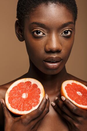Beauty portrait of pretty young half-naked african woman holding grapefruit parts isolated over brown backgroundの写真素材