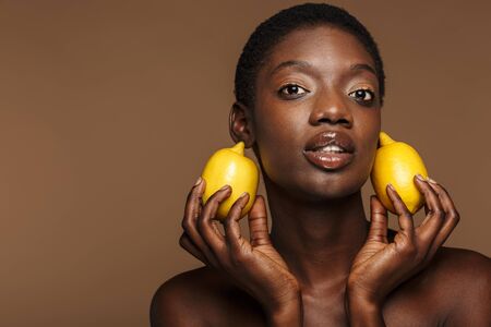 Beauty portrait of pretty young half-naked african woman holding lemons isolated over brown backgroundの写真素材