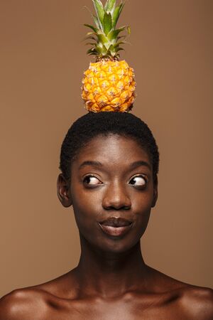 Beauty portrait of pretty young half-naked african woman holding pineapple on her head isolated over brown backgroundの写真素材