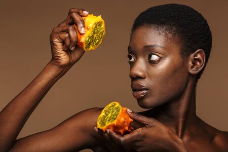 Beauty portrait of pretty young half-naked african woman holding kiwano horned melon isolated over brown backgroundの写真素材