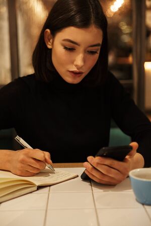 Beautiful young brunette woman sitting at the cafe table indoors, writing on a notebook, holding...