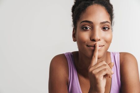 Portrait of african american pleased woman looking at camera while making silence gesture isolated over white backgroundの写真素材