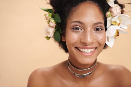 Portrait of young half-naked freckled african american woman with flowers in her hair isolated over beige backgroundの写真素材