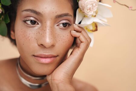 Portrait of young half-naked freckled african american woman with flowers in her hair isolated over beige backgroundの写真素材