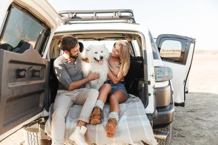 Lovely young happy couple sitting in the back of their car at the beach, playing with dogの写真素材