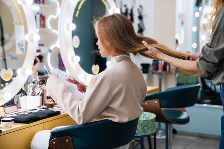 Photo of a concentrated young beautiful woman client reading magazine with hair stylist in saloon.の写真素材