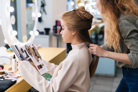 Image of a pretty young beautiful woman client with hair stylist in saloon reading magazine.