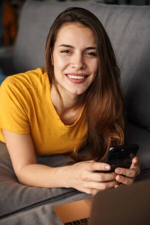 Image of smiling young cute woman using laptop and mobile phone while lying on couch at living roomの写真素材