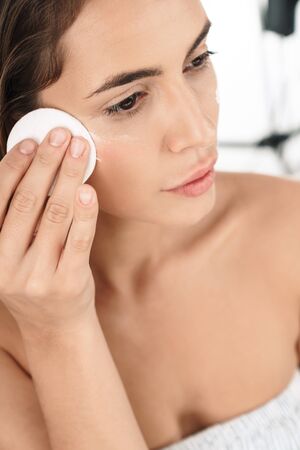 Portrait of adorable young half-naked woman removing makeup with cotton pad isolated over white backgroundの写真素材