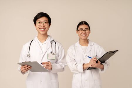 Image of asian young medical doctors in white uniform smiling and holding clipboards isolated over beige backgroundの写真素材