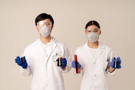Image of asian young doctors wearing uniform and protective masks holding medical tubes with liquid isolated over beige backgroundの写真素材